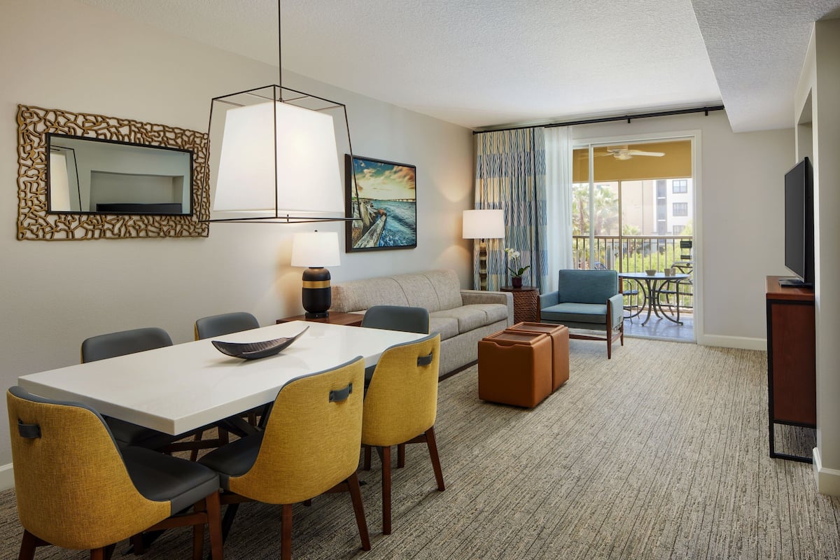 A hotel room with a dining area, a sofa, a balcony with a view, and a TV.