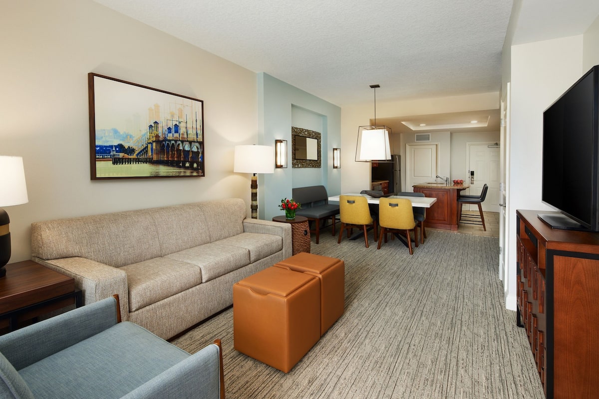 A hotel room with a sofa, ottoman, dining table with chairs, a TV, and a painting on the wall.