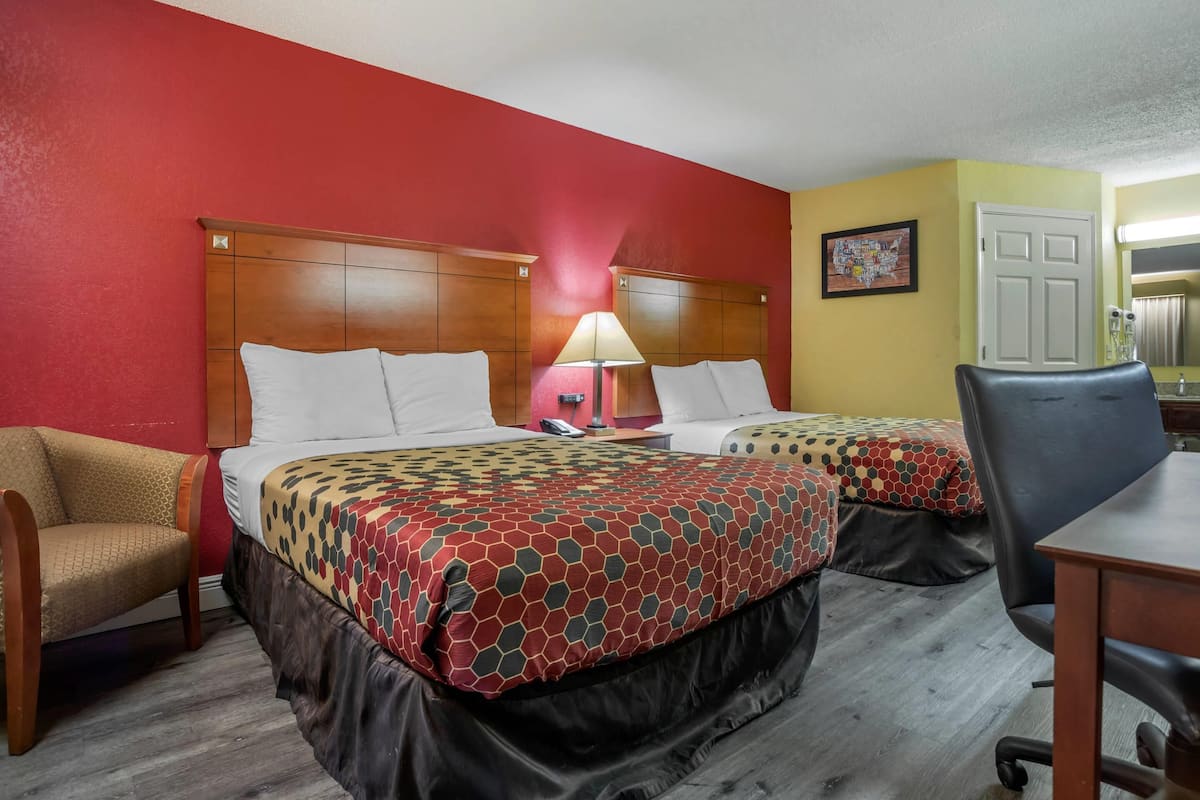 Ein Hotelzimmer mit zwei Betten, einem Holz-Kopfteil, einem Schreibtisch, einem Stuhl, einer Lampe und einer Tür.