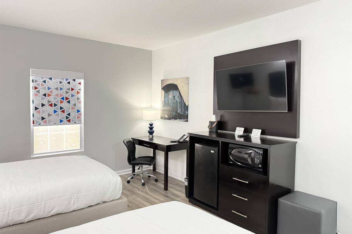 A hotel room with two beds, a desk with a computer, a TV, and a window with a geometric pattern.
