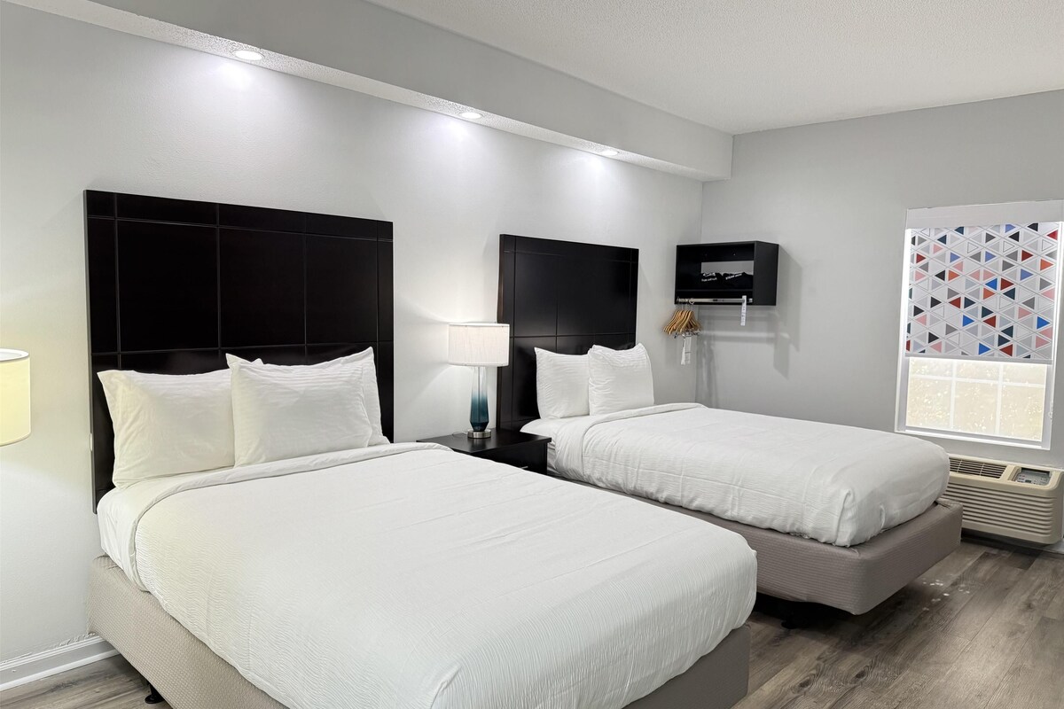 A hotel room with two beds, a TV, a window with geometric curtains, and a wall-mounted heater.