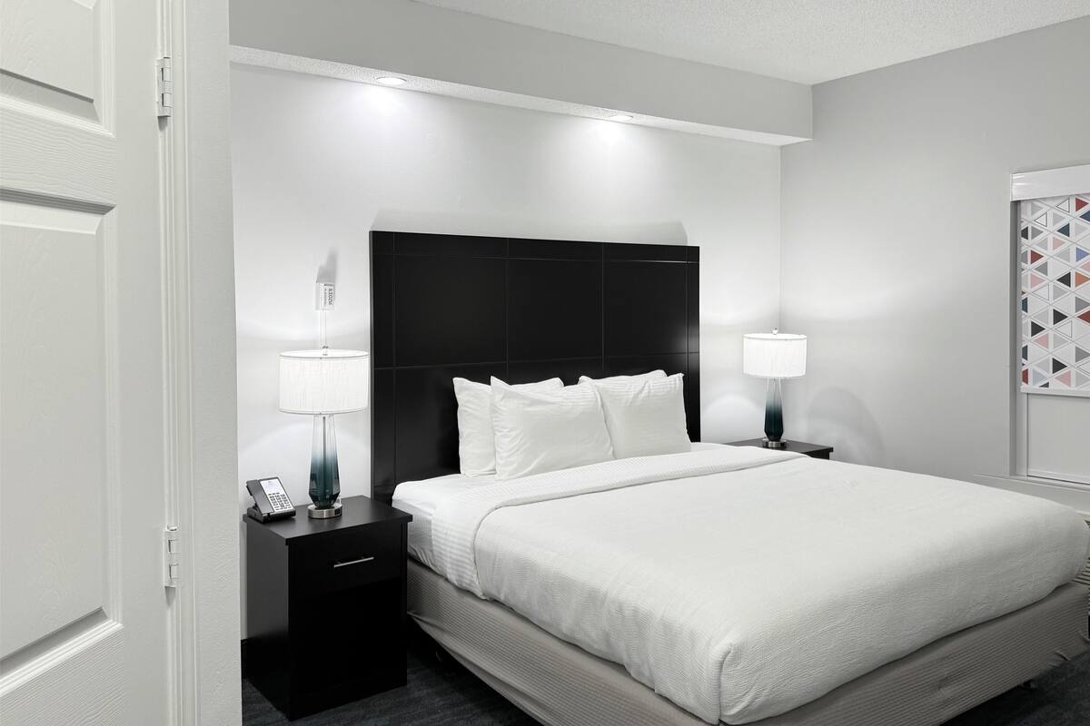 A modern hotel room with a large bed, a black headboard, two bedside tables with lamps, and a small desk with a phone and a lamp.