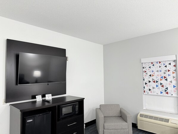 Une chambre d’hôtel dotée d’un téléviseur à écran plat fixé au mur, d’une petite kitchenette équipée d’un four à micro-ondes, d’un fauteuil gris et d’une fenêtre ornée d’un motif géométrique.