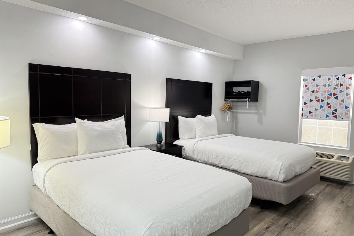 Ein Hotelzimmer mit zwei Betten, einem Fernseher, einem Fenster mit geometrischen Vorhängen und einem Wandheizkörper.