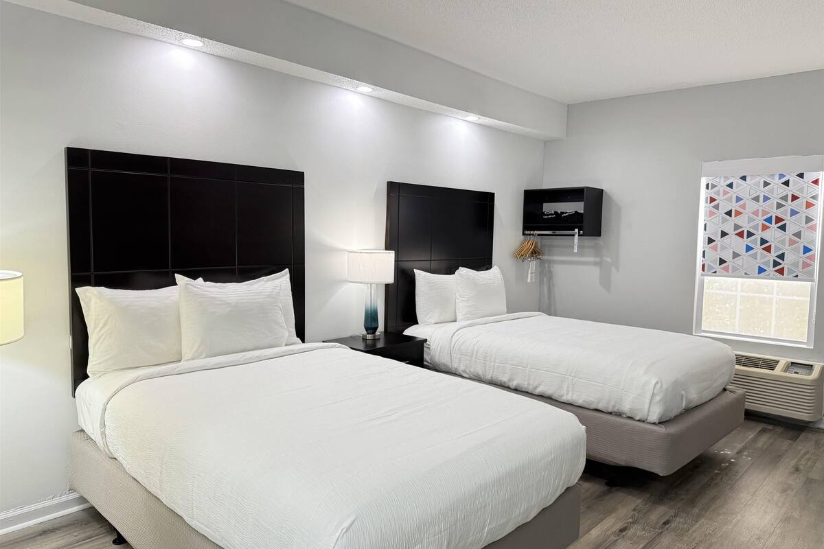 A hotel room with two beds, a TV, a window with geometric curtains, and a wall-mounted heater.