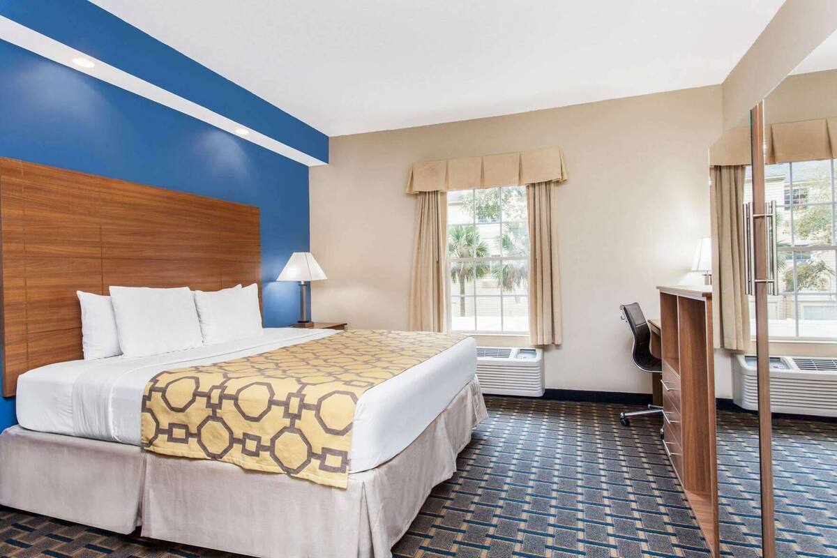 Une chambre d’hôtel comprenant un lit, une table de chevet, une lampe, un appareil de climatisation, un bureau, une chaise et de grandes fenêtres avec des rideaux.
