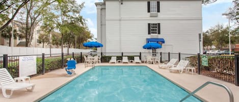 A rectangular swimming pool with a metal ladder, surrounded by white lounge chairs and blue umbrellas.