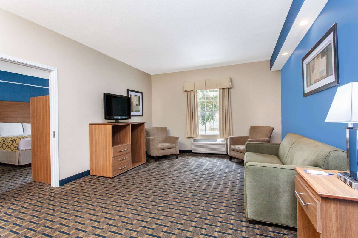 Une chambre d’hôtel avec une moquette à motifs, un téléviseur posé sur un meuble en bois, un canapé, un fauteuil et un bureau avec une lampe.