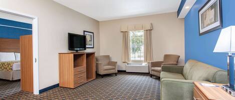 A hotel room with a patterned carpet, a television on a wooden stand, a sofa, a chair, and a desk with a lamp.