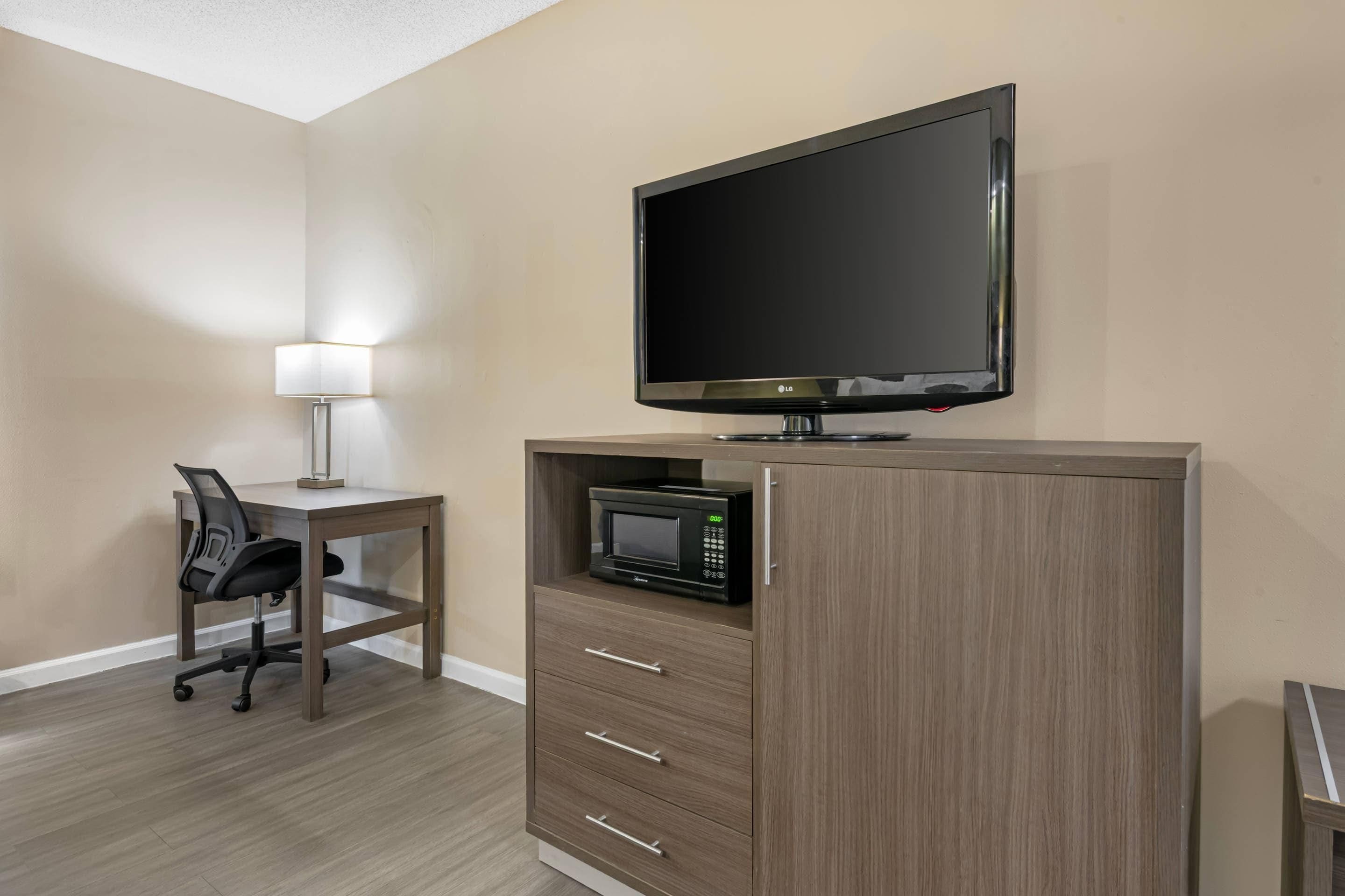 A hotel room with a flat-screen TV, a microwave, a desk with a chair, and a lamp.