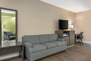 A hotel room with a grey sofa, a desk with a chair, a television, and a cabinet with drawers. - Quality Inn Ormond Beach – Daytona (Ormond Beach)