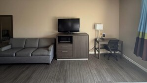 Une chambre d’hôtel avec un canapé gris, un téléviseur posé sur un meuble en bois, un bureau avec une chaise et une lampe.