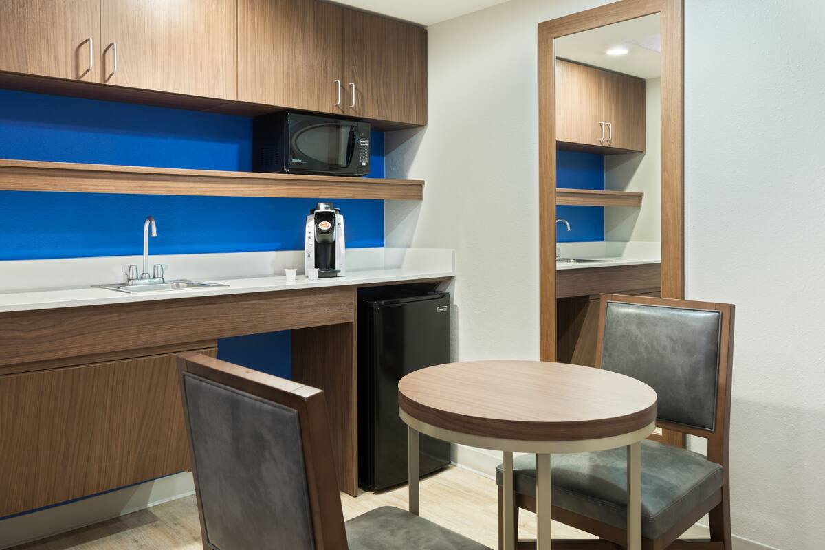 A compact kitchen with wooden cabinets, a microwave, a coffee machine, a sink, and a small refrigerator. There is a round wooden table with two chairs.