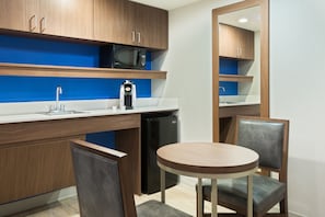 A compact kitchen with wooden cabinets, a microwave, a coffee machine, a sink, and a small refrigerator. There is a round wooden table with two chairs.