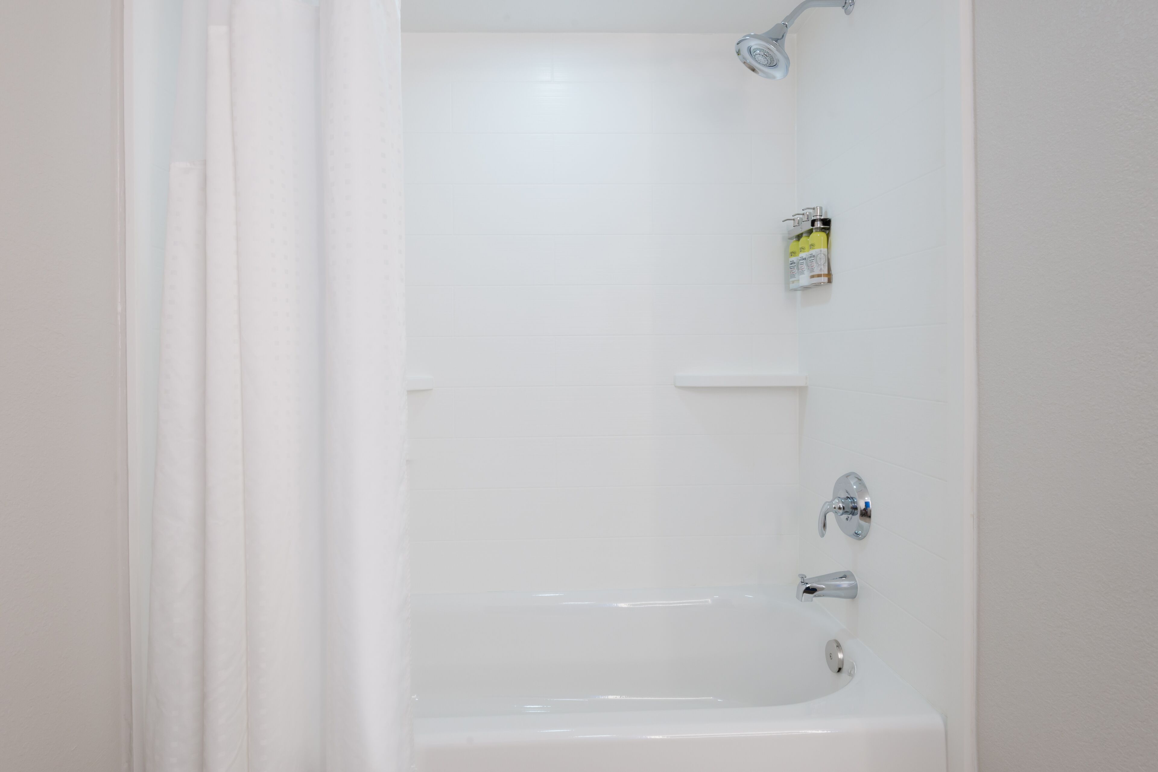 A white bathtub with a showerhead and faucet, a small shelf, and a bottle of shampoo.