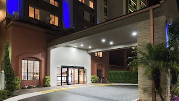 A hotel entrance with a gated driveway, a landscaped area with shrubs and flowers, and a building with illuminated windows.