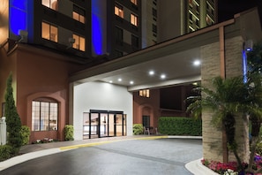 Entrada de hotel con un camino de acceso porticado, un área ajardinada con arbustos y flores, y un edificio con ventanas iluminadas.