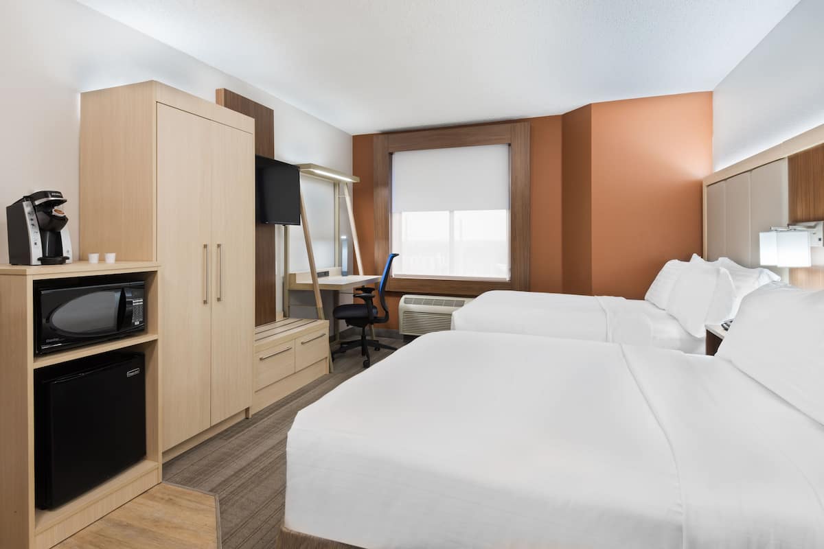 A hotel room with two beds, a desk with a chair, a microwave, a refrigerator, a TV, and a window with blinds.
