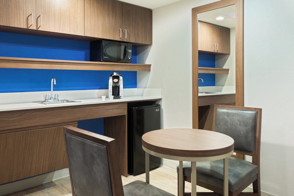 A compact kitchen with a microwave, coffee maker, and refrigerator. There is a round table with two chairs and a mirror above the sink.