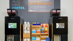 A coffee station with two coffee machines, a screen displaying a man playing guitar, and shelves with coffee cups and boxes. - Holiday Inn Express & Suites Nearest Universal Orlando by IHG (Orlando)