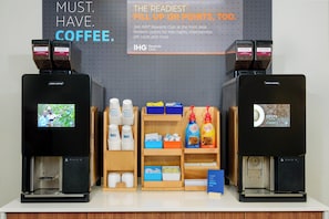 A coffee station with two coffee machines, a screen displaying a man playing guitar, and shelves with coffee cups and boxes.
