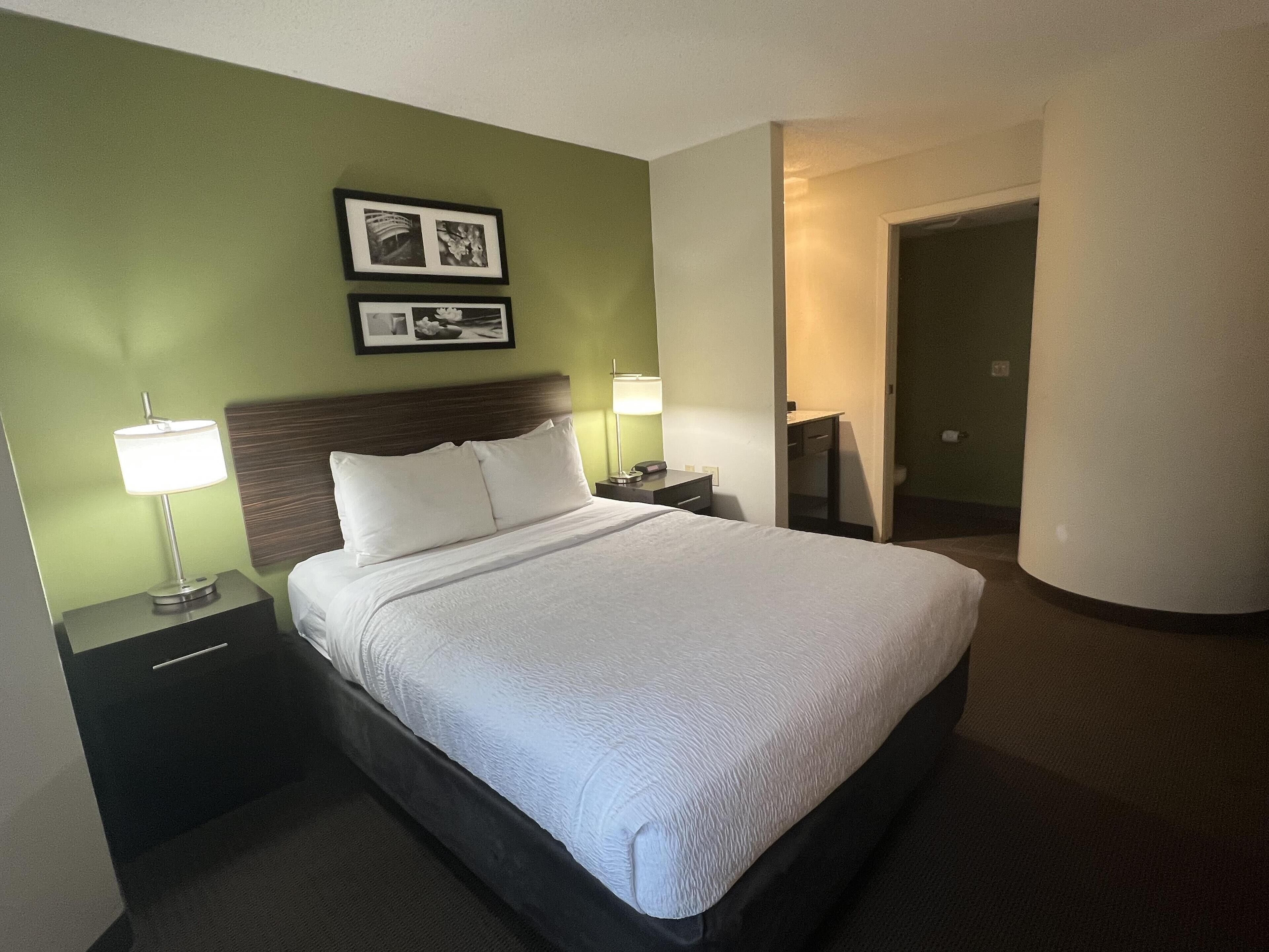 A hotel room with a large bed, two bedside tables with lamps, a headboard, a framed picture on the wall, and a door leading to another room.
