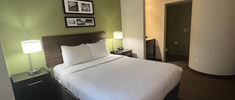 A hotel room with a large bed, two bedside tables with lamps, a headboard, a framed picture on the wall, and a door leading to another room.