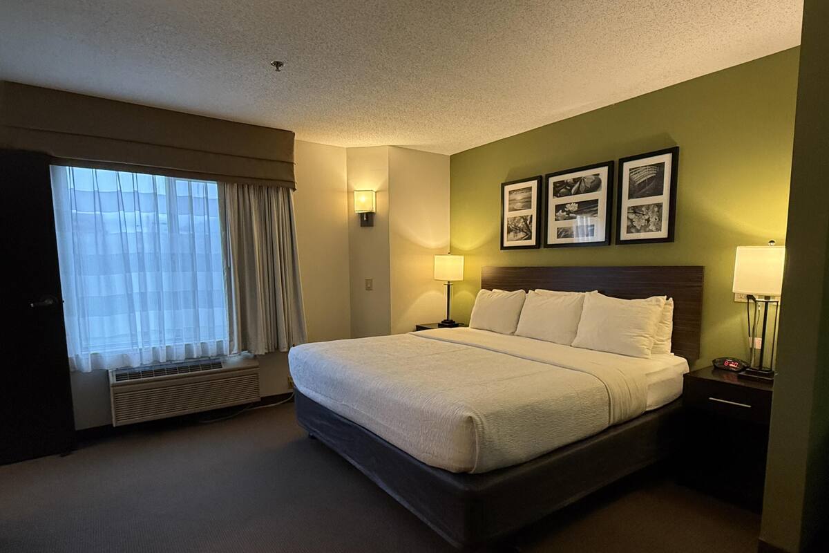 A hotel room with a large bed, a window with curtains, a wall-mounted air conditioner, and framed pictures above the bed.