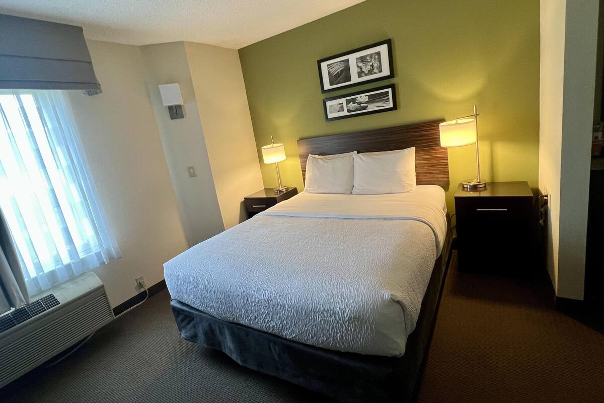 A hotel room with a bed, two bedside tables with lamps, a window with curtains, and a wall-mounted picture.