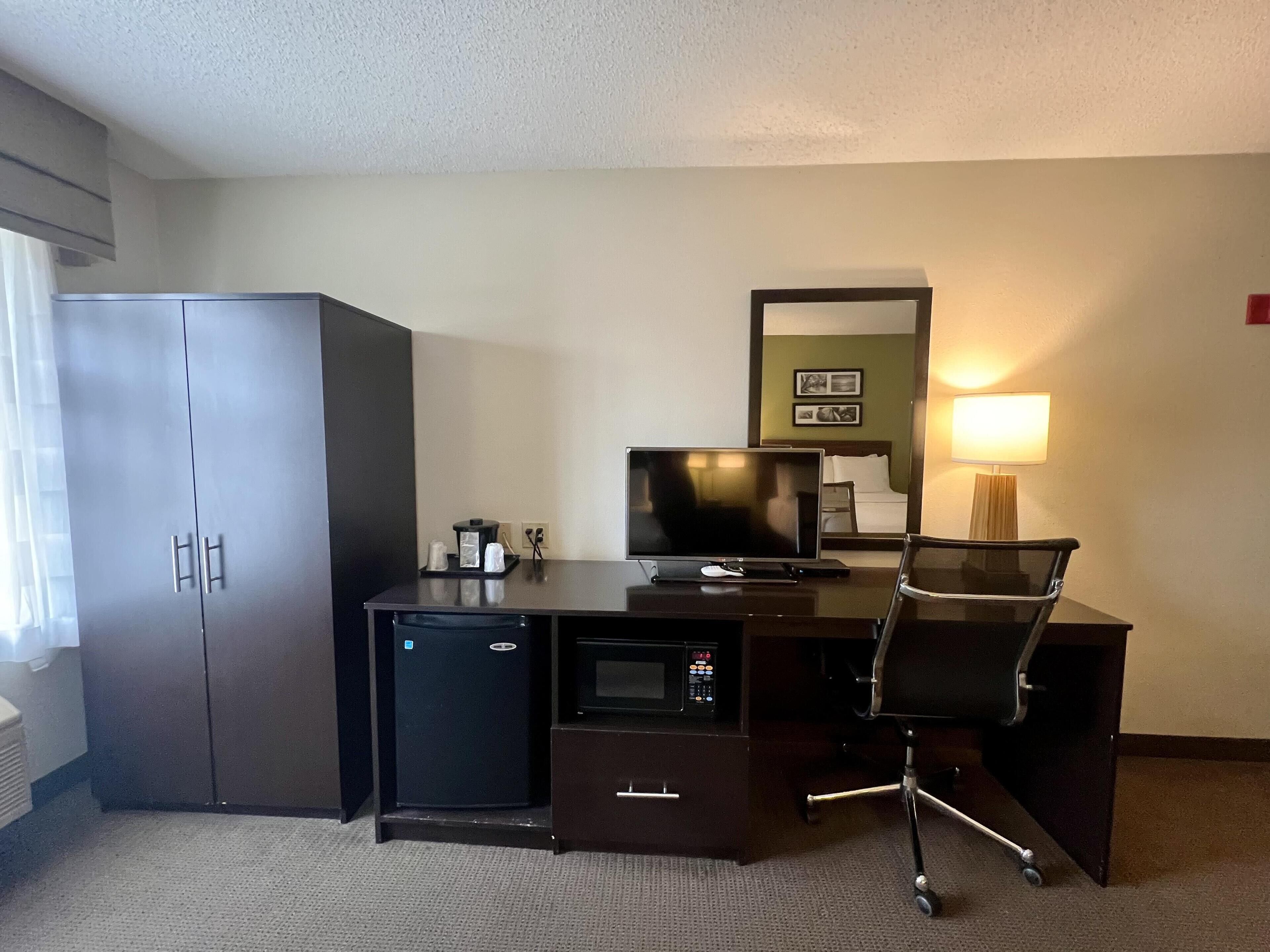 A hotel room with a dark wooden desk, a small refrigerator, a television, a chair, and a bed with a nightstand.