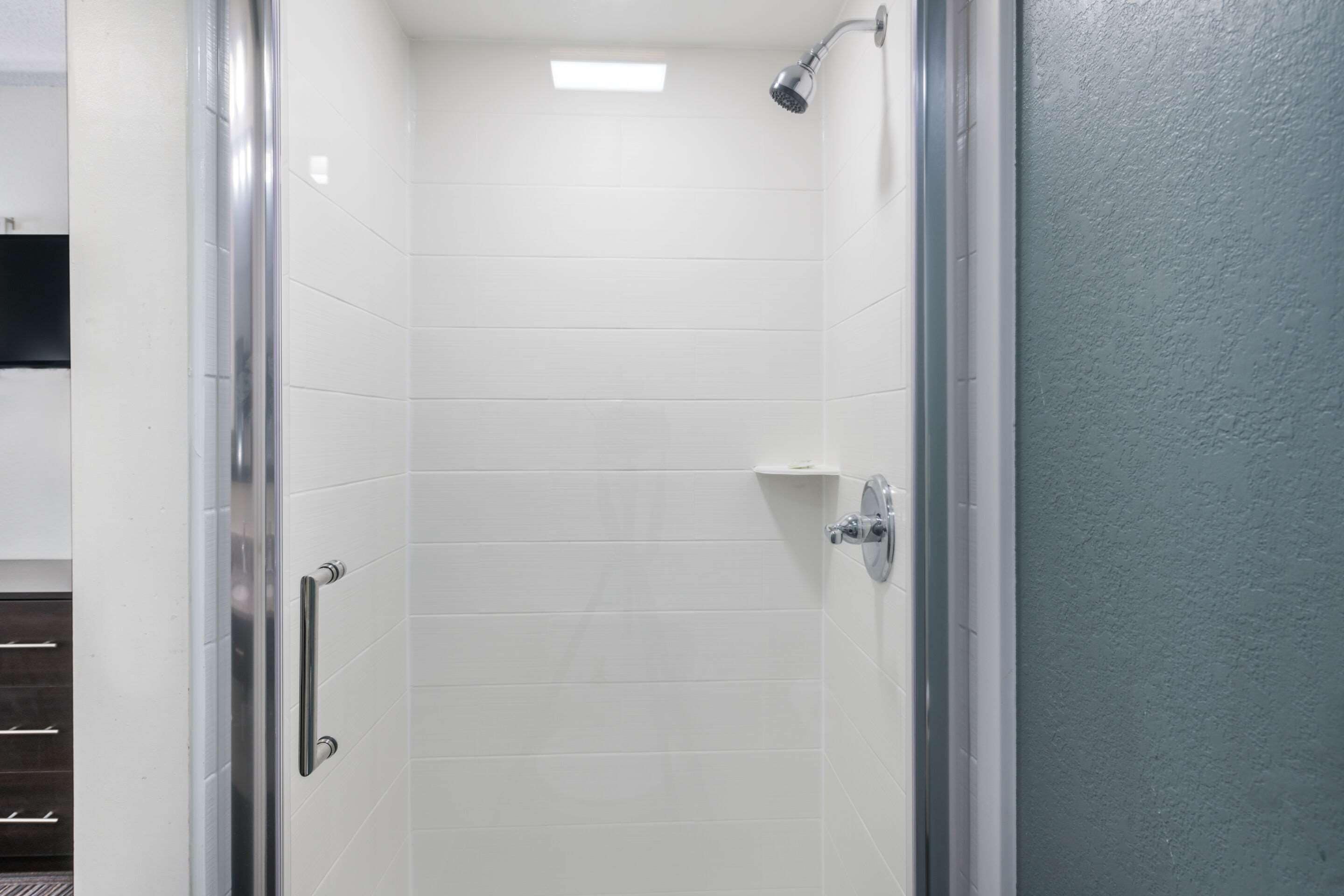 A shower with a glass door, white tiles, and a small shelf.