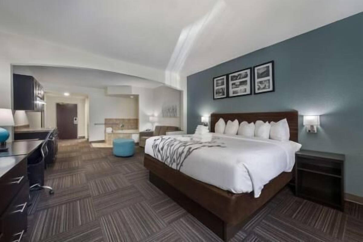 A hotel room with a large bed, a desk, a chair, a blue ottoman, and a bathroom visible in the background.