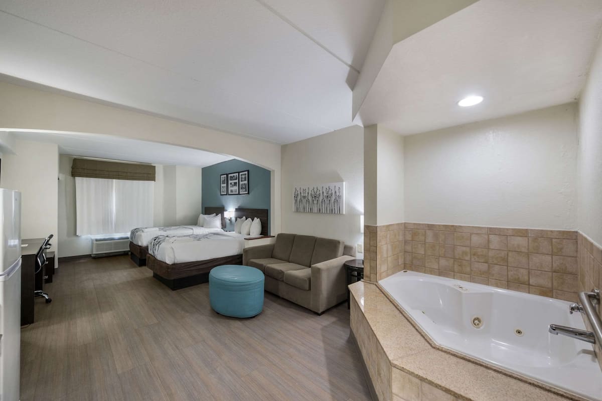 A hotel room with a bed, a sofa, a blue ottoman, a bathroom with a bathtub, and a desk with a chair.