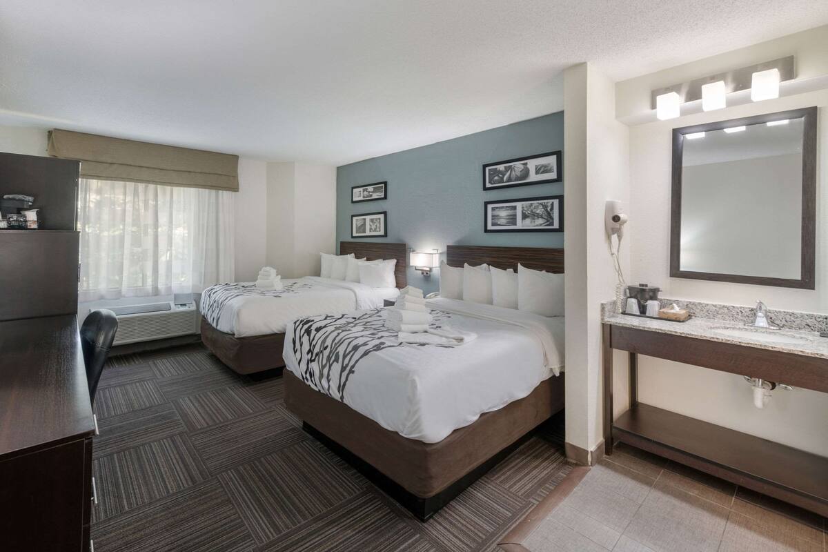 A hotel room with two beds, a desk, a dresser, a vanity with a mirror, and framed pictures on the wall.