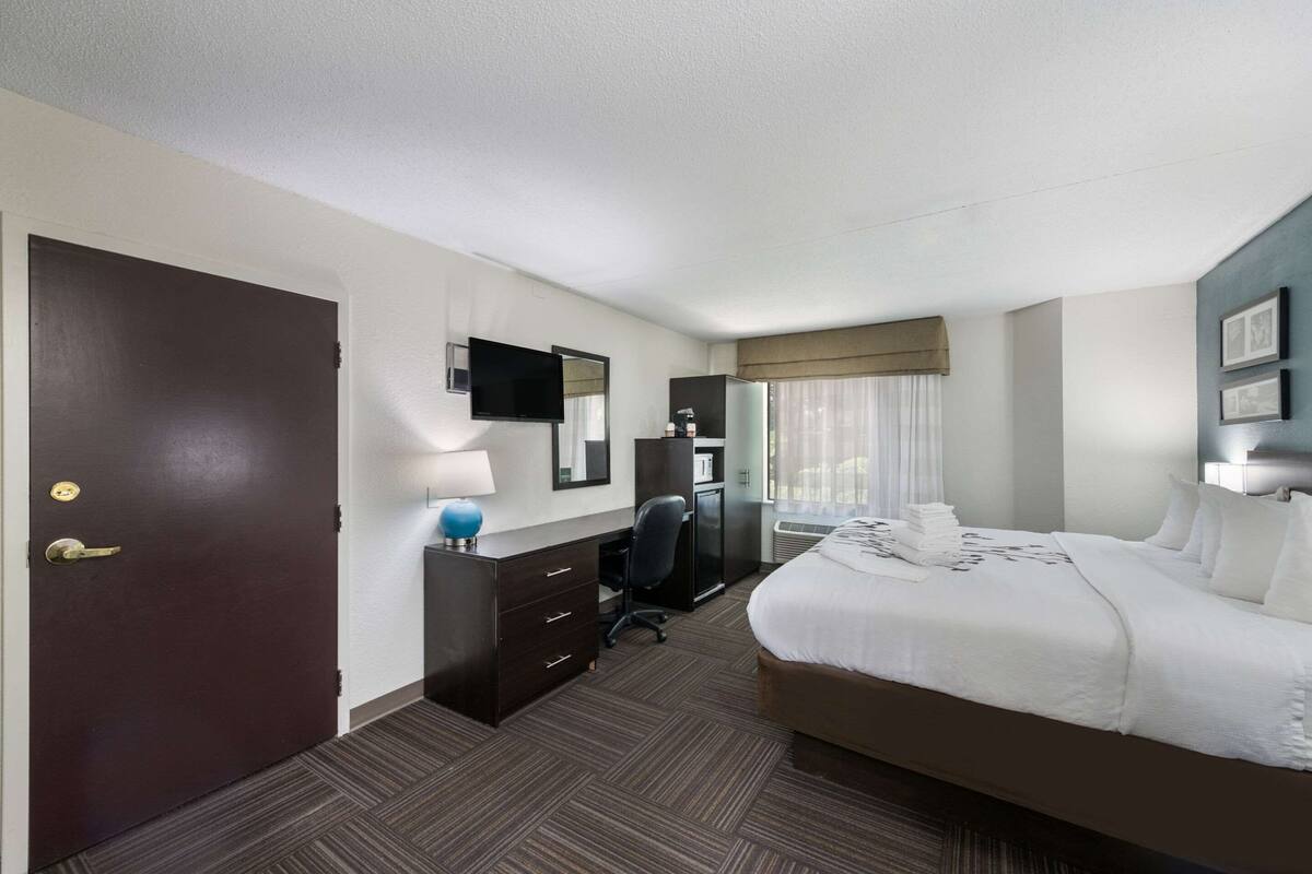 A hotel room with a large bed, a desk with a chair, a TV, a wardrobe, and a window with curtains.