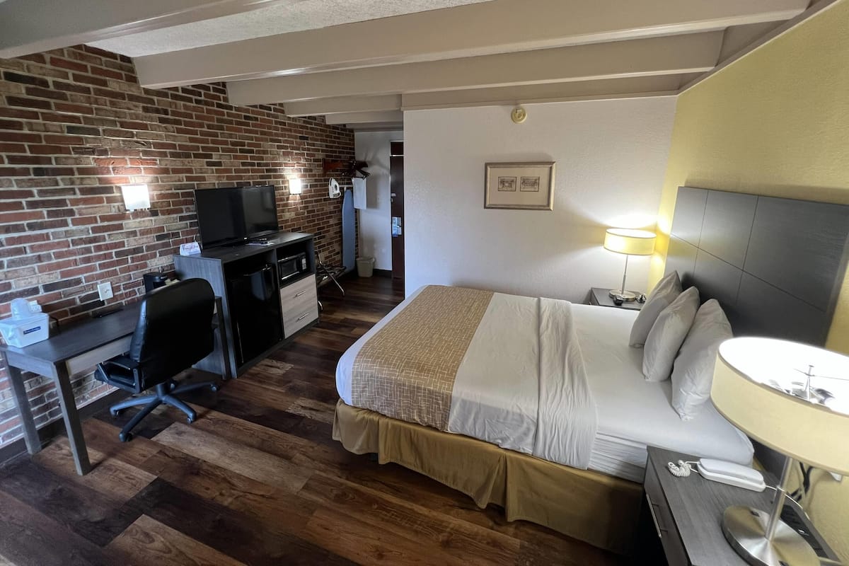 A hotel room with a brick wall, a desk with a television, a bed with a headboard, and a bedside table with a lamp.