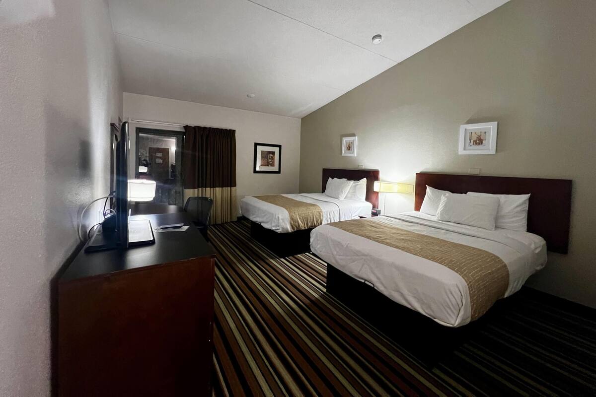 A hotel room with two beds, a desk, a chair, and a view of the hallway.