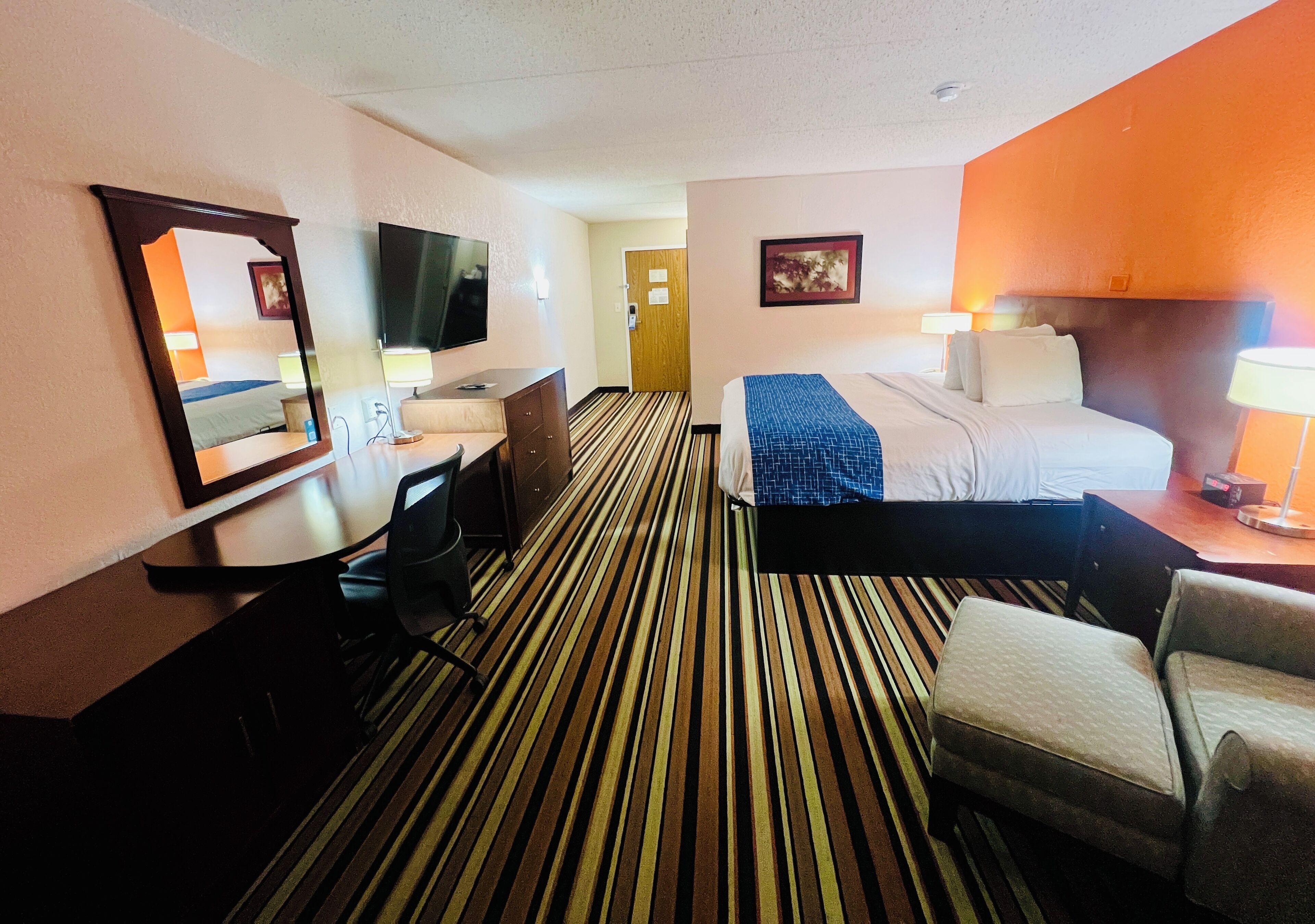 A hotel room with a striped carpet, a bed with a blue blanket, a desk with a chair, a television, and a door leading to another room.