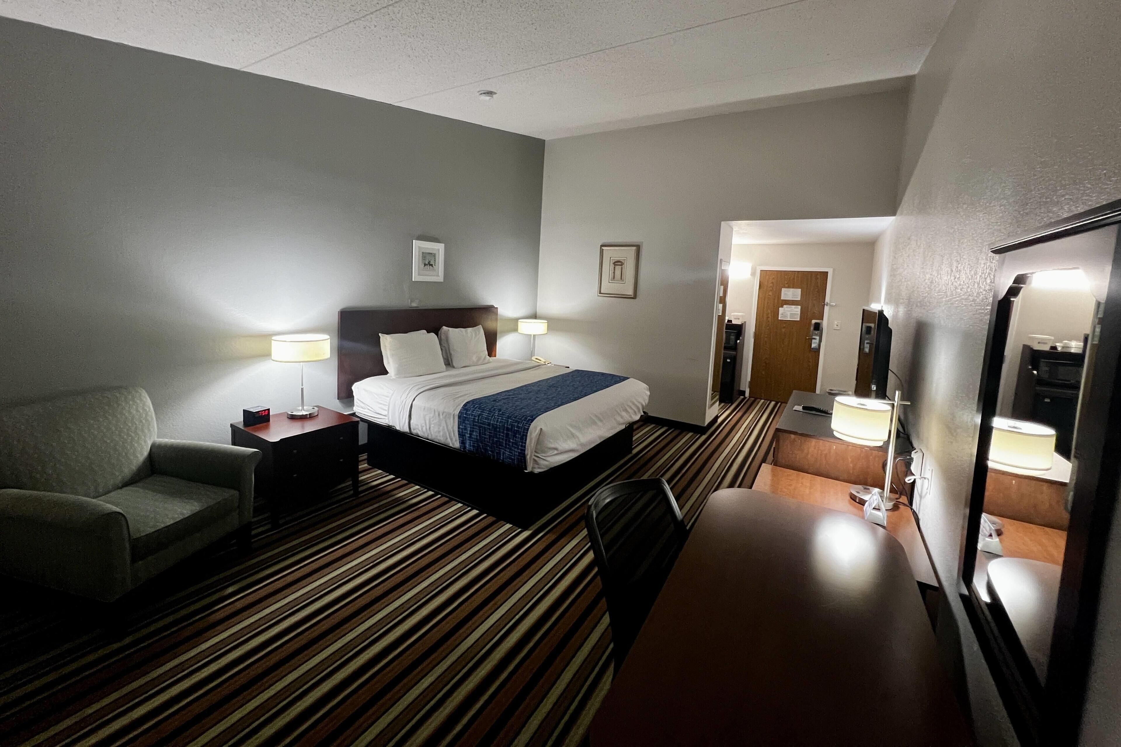 Une chambre d’hôtel comprenant un lit, des tables de chevet, une chaise, un bureau, un miroir et une porte donnant sur une autre pièce.
