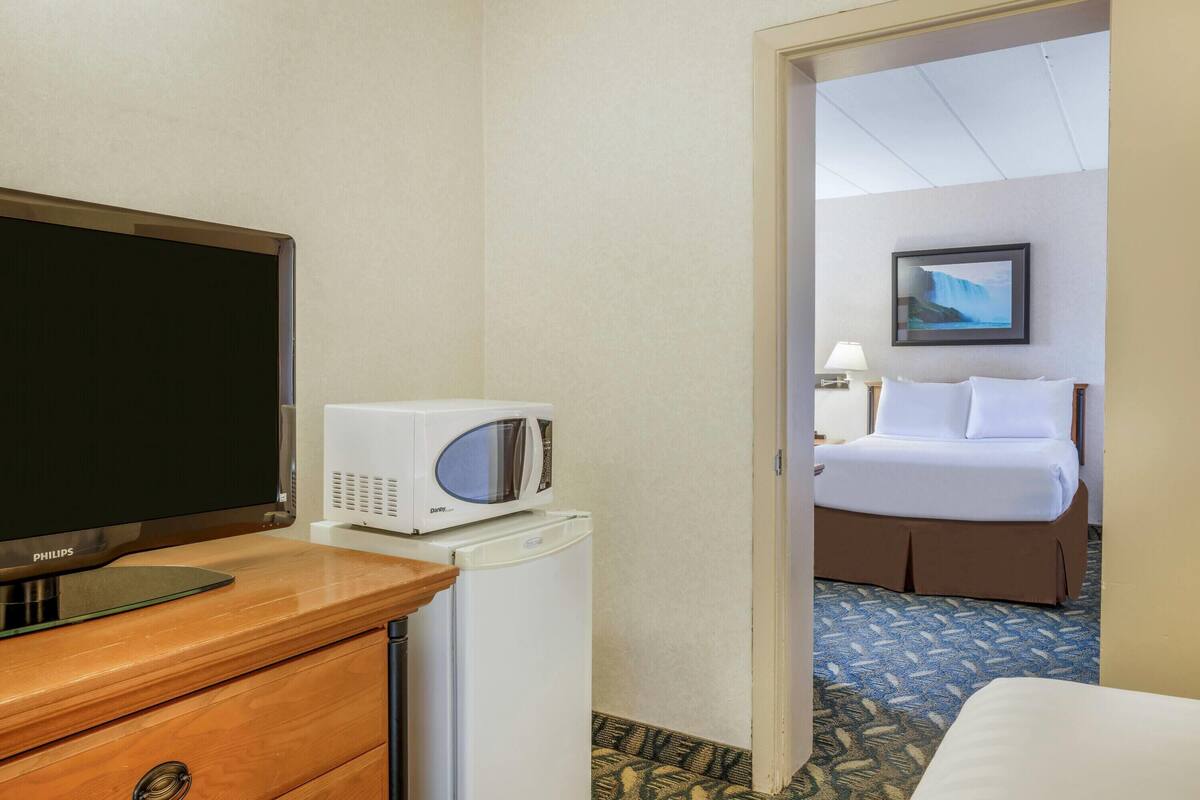 Habitación de hotel con televisión, microondas, refrigerador, cama y mesita de noche con lámpara.