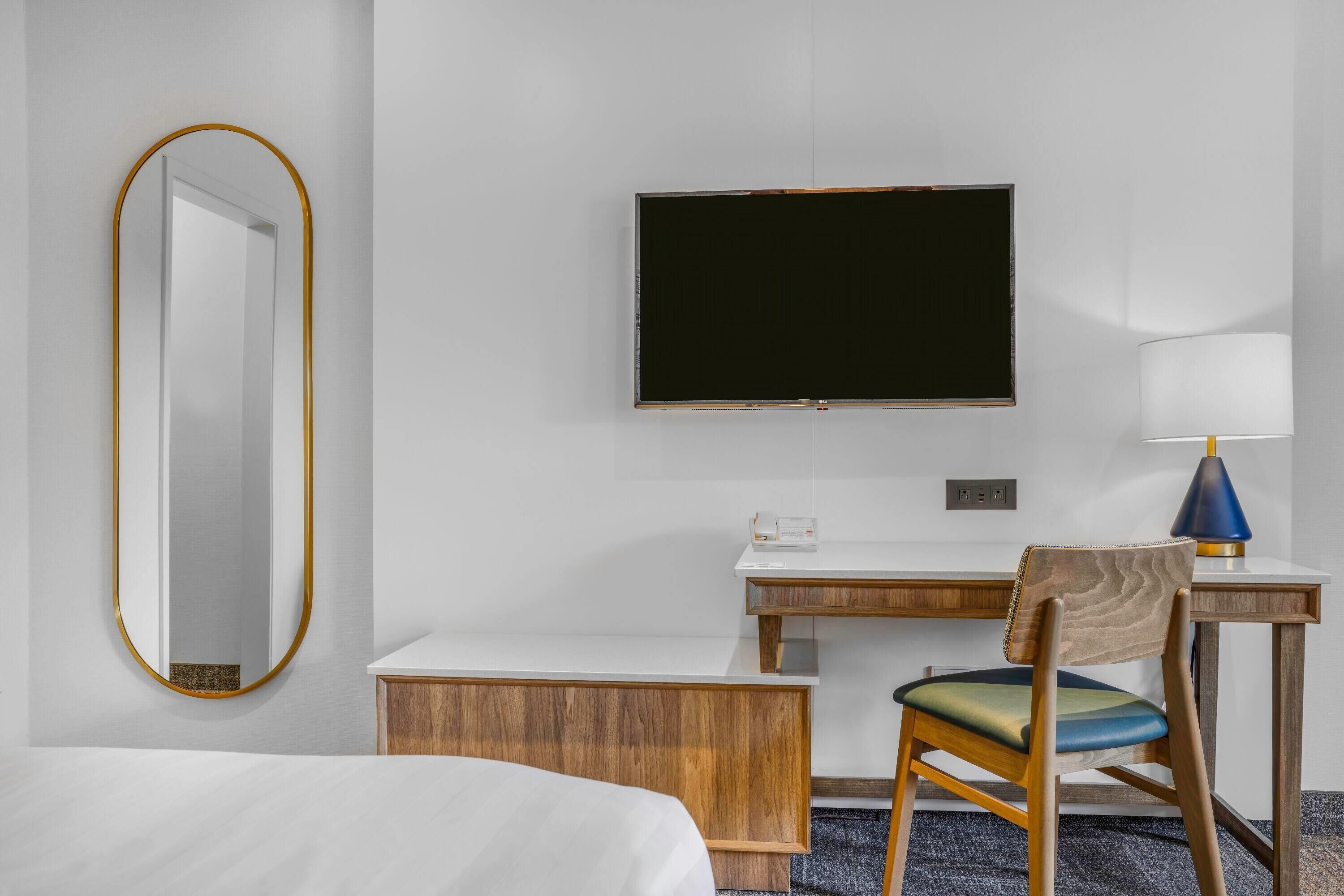 Quarto de hotel com cama, escrivaninha com cadeira, televisão de tela plana, abajur e espelho.