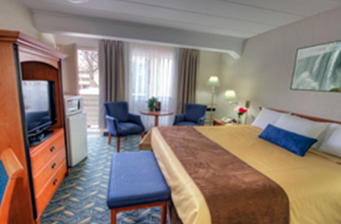 A hotel room with a bed, a television, two chairs, a small table, and a window with curtains.