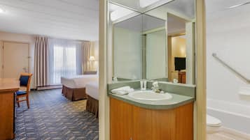 Una habitación de hotel con cama, escritorio, baño con lavamanos y espejo, y ducha.