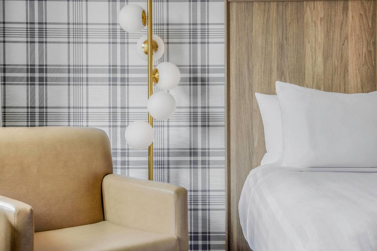 Una habitación de hotel con papel tapiz escocés, un sillón color beige, un cabecero de madera y una cama blanca.