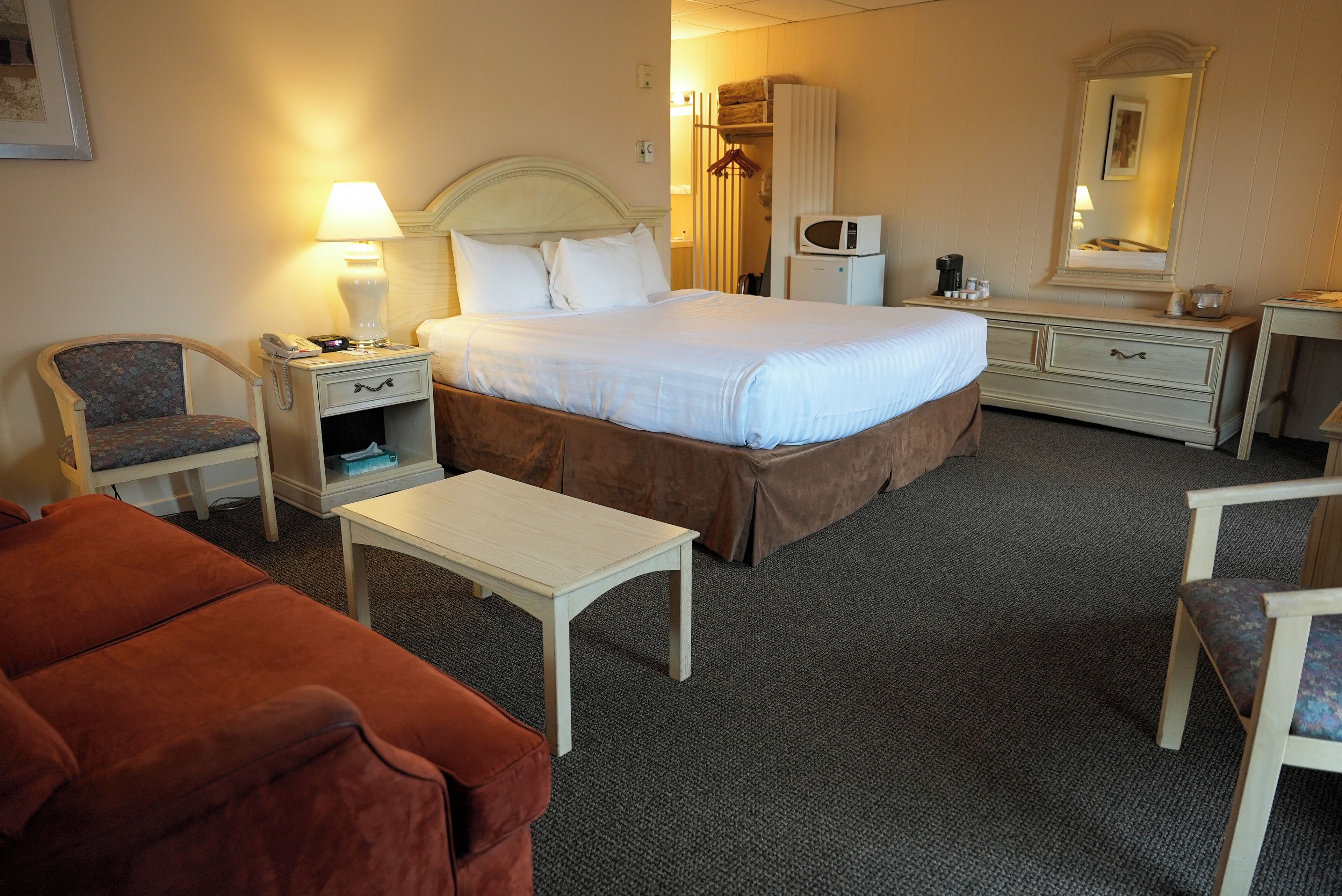 A hotel room with a large bed, a bedside table with a lamp, a chair, a small table, a dresser, and a microwave.