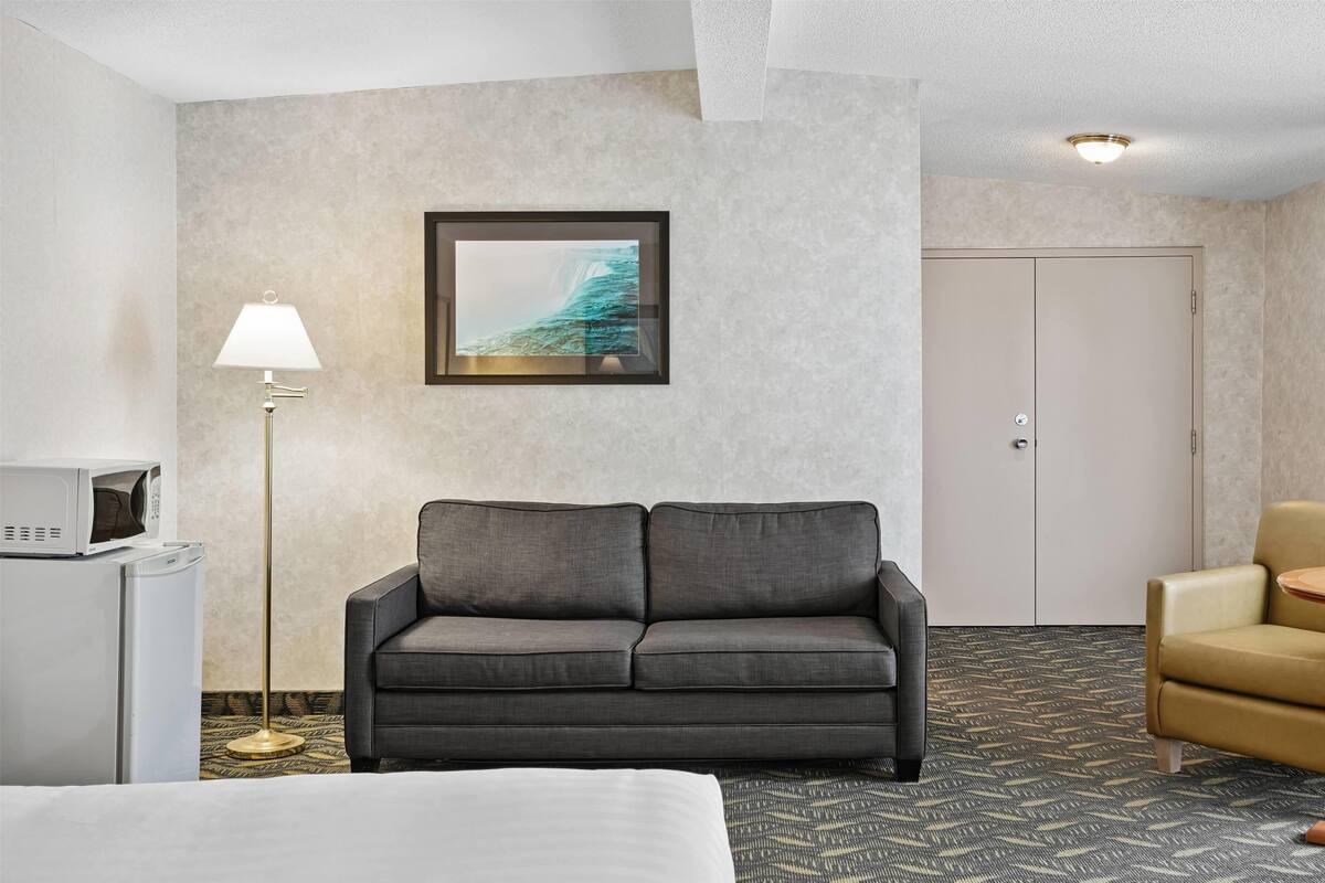 Habitación de hotel con un sofá gris, una mesita redonda pequeña, una lámpara y un cuadro en la pared.