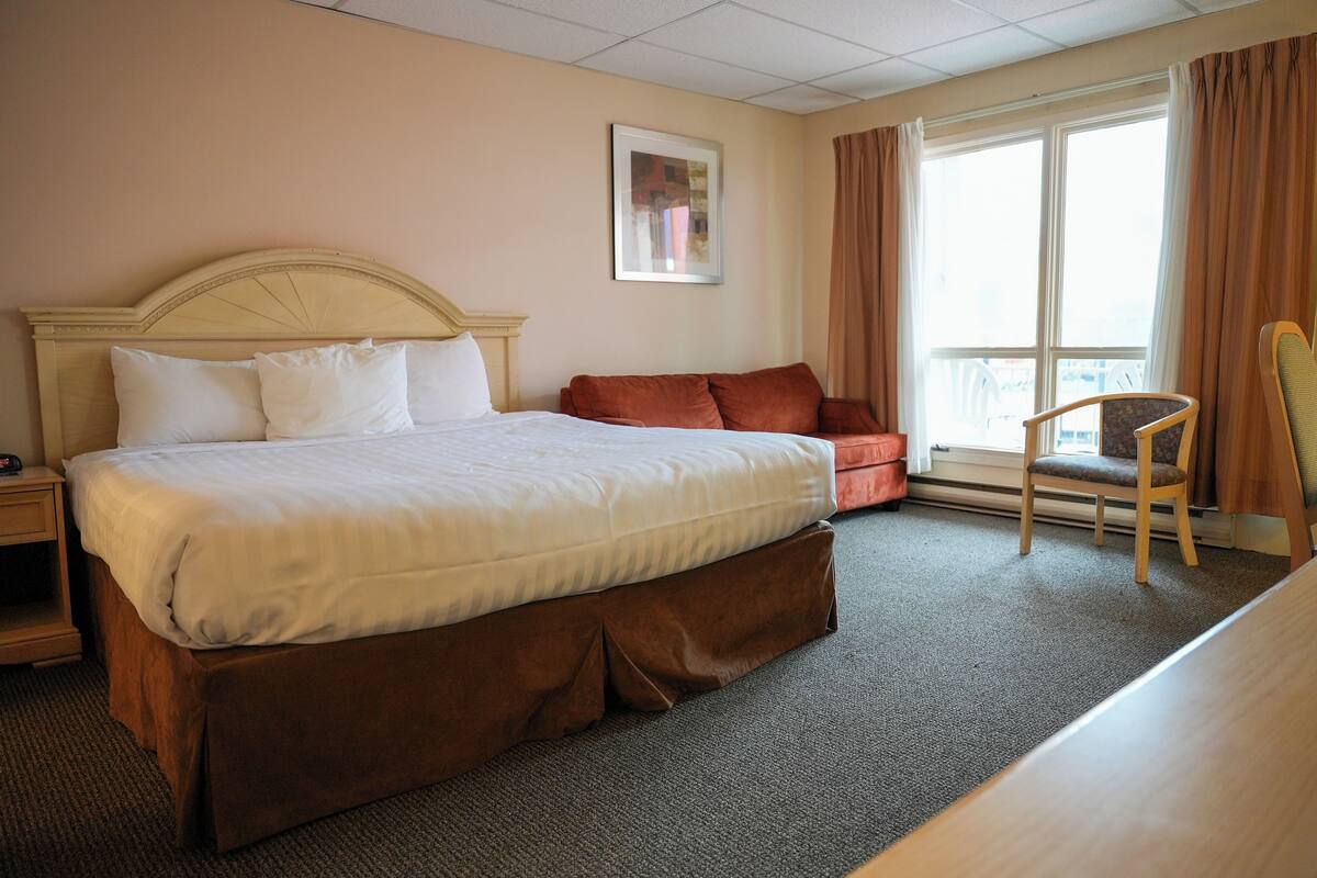 Habitación de hotel con una cama grande, una mesita de noche de madera, una silla y un ventanal con cortinas.