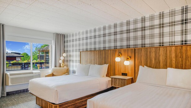 Habitación de hotel con dos camas, un ventanal con vistas a un edificio y zonas verdes, y una pared con estampado escocés.