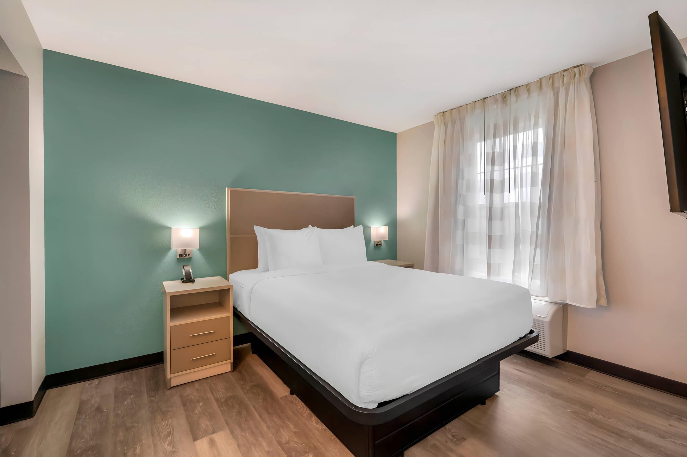 A hotel room with a bed, bedside tables, a nightstand, a window with curtains, and a wall-mounted TV.