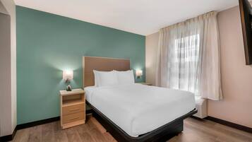 A hotel room with a bed, bedside tables, a nightstand, a window with curtains, and a wall-mounted TV.